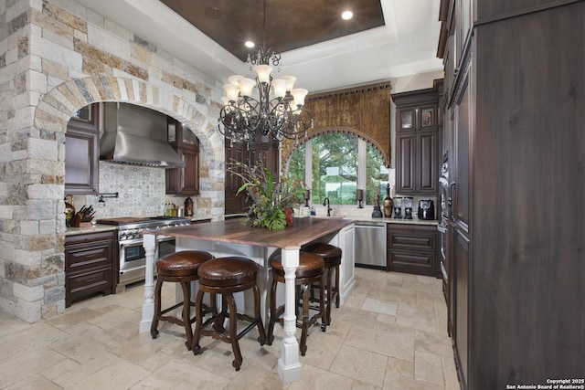 2219 Clubs Dr, Boerne, TX 78006 - kitchen featuring wall chimney range hood, stainless steel appliances, stone tile flooring, tasteful backsplash, and a raised ceiling