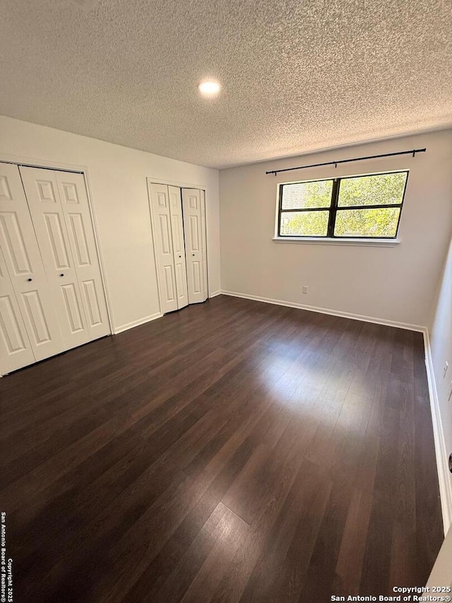 unfurnished bedroom featuring a textured ceiling, two closets, dark wood finished floors, baseboards, and recessed lighting