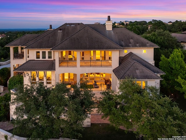 52 Oakland Hls, Boerne, TX 78006 - back of house at dusk featuring a balcony, a chimney, a tiled roof, stucco siding, and a patio