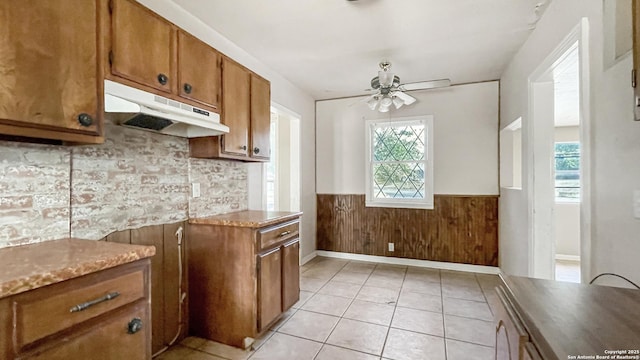 103 Pollydale Ave, San Antonio, TX 78223 - kitchen with under cabinet range hood, brown cabinetry, a wainscoted wall, a ceiling fan, and light tile patterned floors
