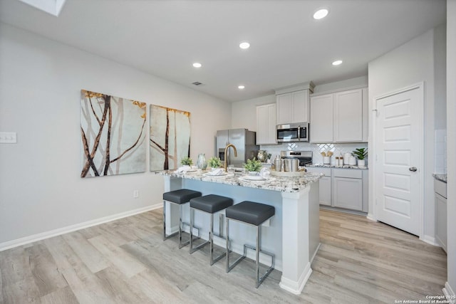 11212 Schmidt Ln, Converse, TX 78109 - kitchen featuring appliances with stainless steel finishes, light wood-style flooring, a kitchen bar, an island with sink, and light stone countertops