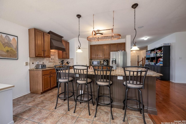 4706 Granite Shls, San Antonio, TX 78244 - kitchen with stainless steel appliances, custom range hood, brown cabinets, backsplash, and a breakfast bar