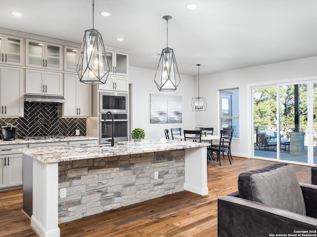 555 Mystic Pkwy, Spring Branch, TX 78070 - kitchen featuring appliances with stainless steel finishes, wood finished floors, light stone countertops, decorative backsplash, and an island with sink
