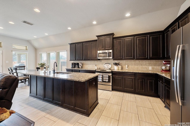 2129 Rustling Way, Seguin, TX 78155 - kitchen featuring appliances with stainless steel finishes, lofted ceiling, decorative backsplash, light stone counters, and a center island with sink