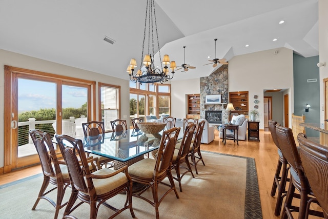 dining room with high vaulted ceiling, a stone fireplace, light wood-style flooring, ceiling fan with notable chandelier, and visible vents