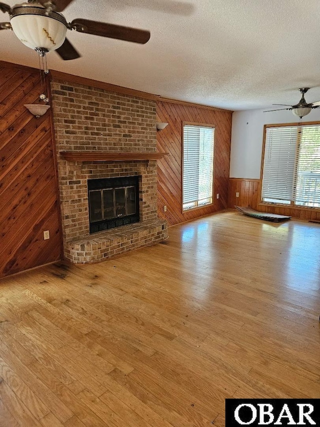 unfurnished living room featuring a fireplace, light wood finished floors, a ceiling fan, wooden walls, and a textured ceiling