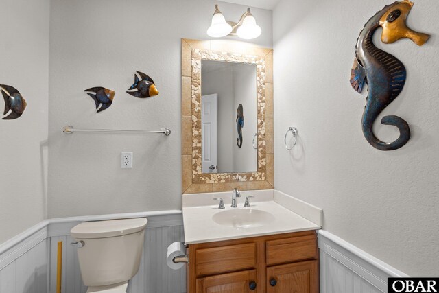 half bathroom featuring vanity, wainscoting, toilet, and a decorative wall