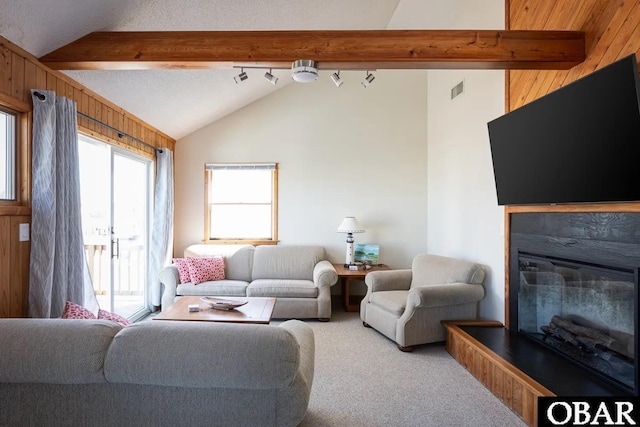 carpeted living room with visible vents, lofted ceiling with beams, a glass covered fireplace, wood walls, and track lighting