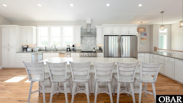 kitchen with stainless steel appliances, white cabinets, hanging light fixtures, and wall chimney range hood