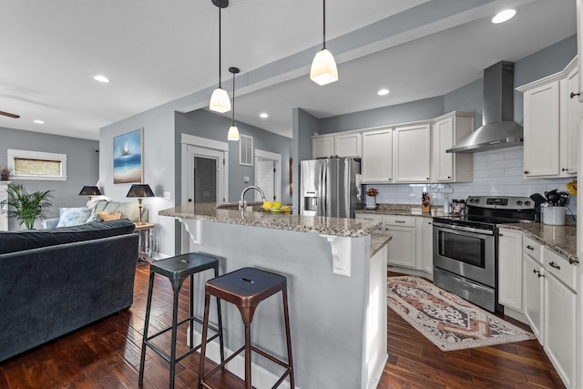 kitchen with an island with sink, open floor plan, stainless steel appliances, wall chimney range hood, and white cabinetry