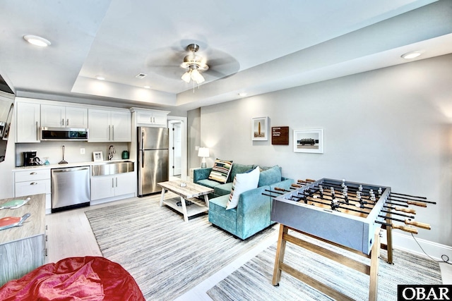 living room featuring a ceiling fan, baseboards, a tray ceiling, recessed lighting, and light wood-type flooring