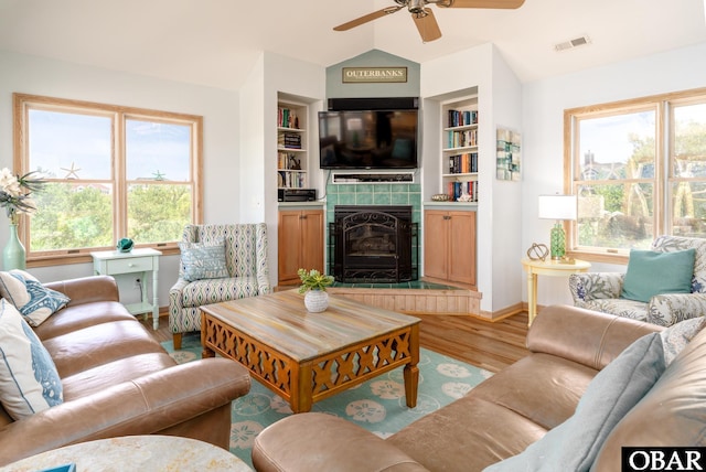living area with a ceiling fan, wood finished floors, vaulted ceiling, a tile fireplace, and visible vents