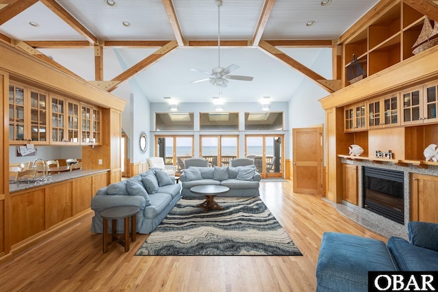 living area with a ceiling fan, a glass covered fireplace, light wood-style flooring, high vaulted ceiling, and beam ceiling