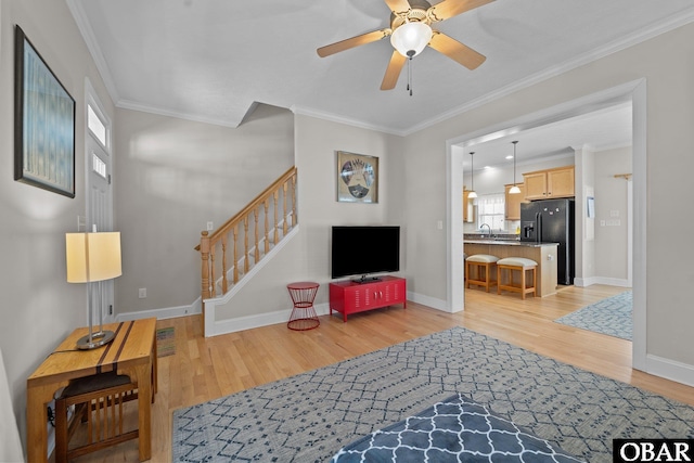 living area with ceiling fan, light wood finished floors, baseboards, ornamental molding, and stairway