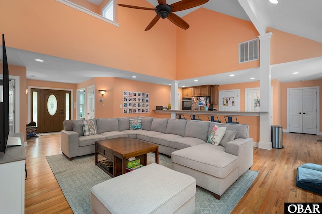 living area with high vaulted ceiling, light wood-style flooring, a ceiling fan, visible vents, and decorative columns