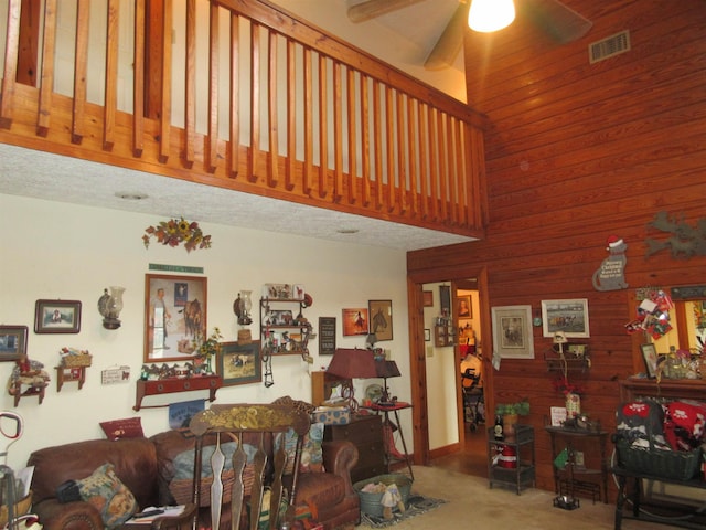 living area with visible vents, a ceiling fan, a towering ceiling, carpet flooring, and wood walls