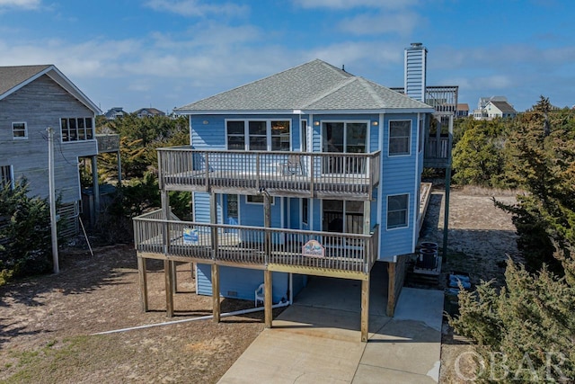 rear view of property with a balcony, a shingled roof, concrete driveway, a wooden deck, and a chimney