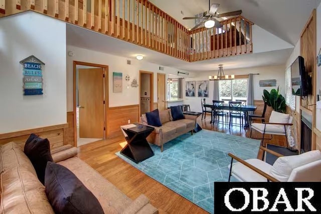 living room with a wainscoted wall, ceiling fan with notable chandelier, a high ceiling, wood walls, and wood finished floors