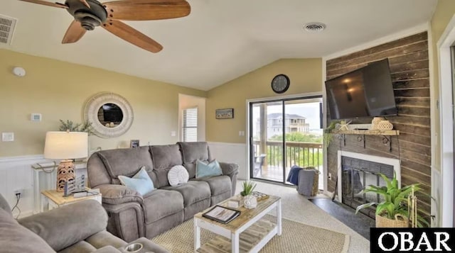 living room with a large fireplace, visible vents, a ceiling fan, a wainscoted wall, and vaulted ceiling