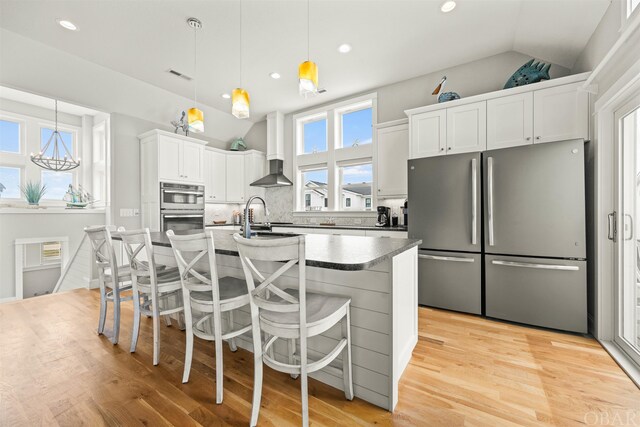 kitchen featuring decorative light fixtures, lofted ceiling, appliances with stainless steel finishes, white cabinetry, and wall chimney range hood