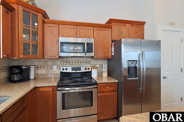 kitchen with appliances with stainless steel finishes, brown cabinets, glass insert cabinets, and light stone countertops