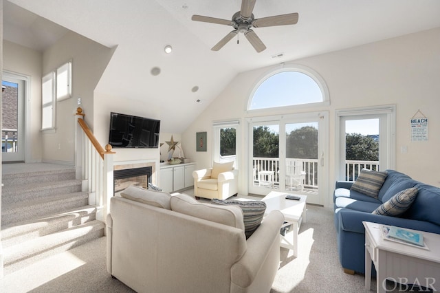 living room featuring light colored carpet, stairway, a tiled fireplace, a ceiling fan, and high vaulted ceiling