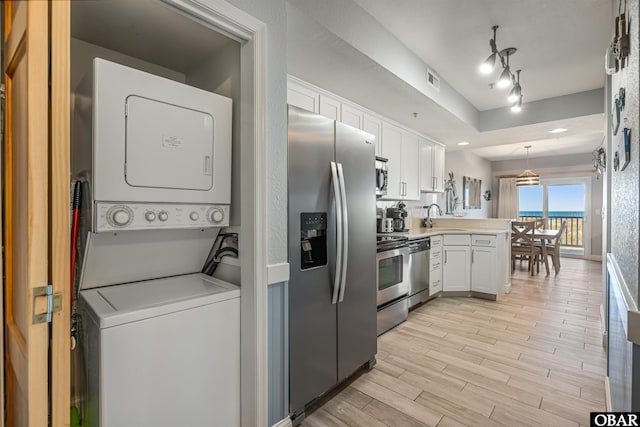 kitchen featuring appliances with stainless steel finishes, hanging light fixtures, light countertops, stacked washing maching and dryer, and white cabinetry