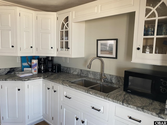 kitchen with glass insert cabinets, black microwave, dark stone counters, white cabinetry, and a sink