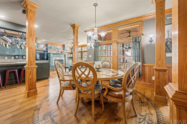 dining space featuring a wainscoted wall, light wood-style flooring, ornate columns, a fireplace, and a chandelier