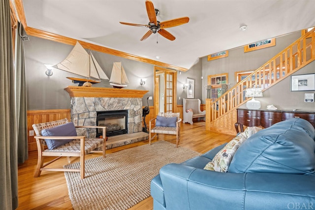 living room featuring light wood-style flooring, stairway, wainscoting, ceiling fan, and a stone fireplace