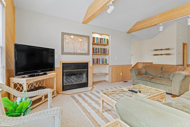 living room with built in features, light colored carpet, lofted ceiling with beams, a glass covered fireplace, and wainscoting