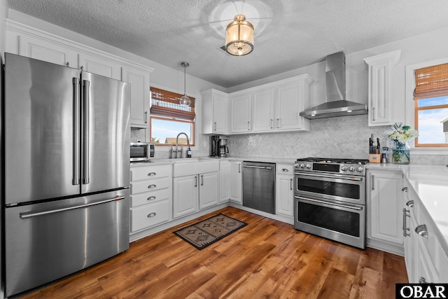 kitchen featuring a sink, wall chimney exhaust hood, stainless steel appliances, white cabinetry, and wood finished floors