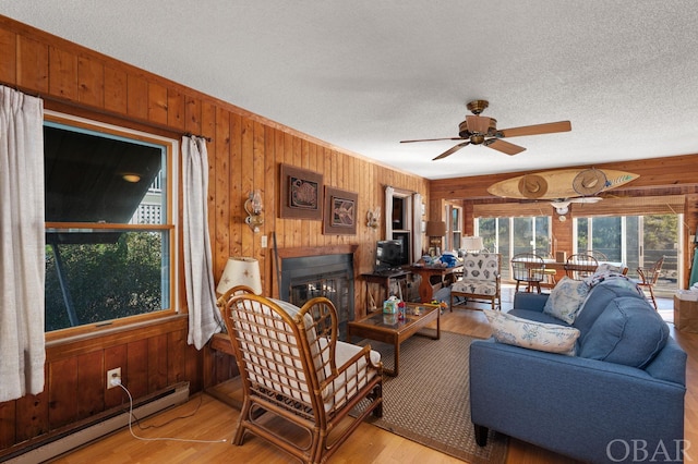 living area featuring a baseboard radiator, light wood-style floors, a glass covered fireplace, wood walls, and a textured ceiling