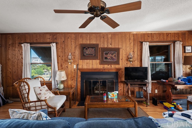 living room with wooden walls, a ceiling fan, a glass covered fireplace, wood finished floors, and a textured ceiling