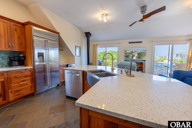 kitchen with light stone counters, appliances with stainless steel finishes, a kitchen island with sink, and brown cabinets