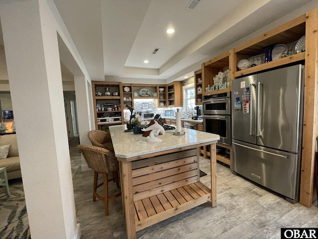 kitchen with light stone counters, open shelves, stainless steel appliances, a raised ceiling, and brown cabinetry