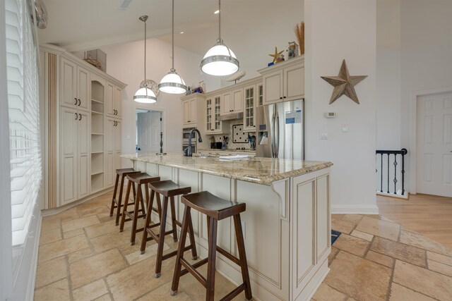 kitchen with stone tile floors, stainless steel fridge with ice dispenser, glass insert cabinets, cream cabinetry, and pendant lighting