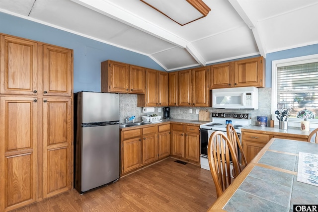 kitchen featuring lofted ceiling with beams, white appliances, a sink, wood finished floors, and brown cabinets