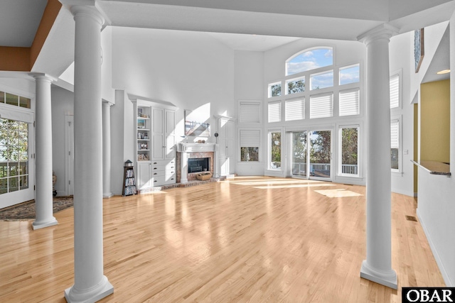 unfurnished living room with a fireplace with raised hearth, light wood-type flooring, a towering ceiling, and decorative columns