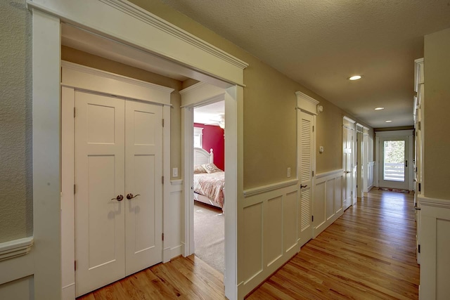 corridor with a textured ceiling, light wood finished floors, wainscoting, and a decorative wall