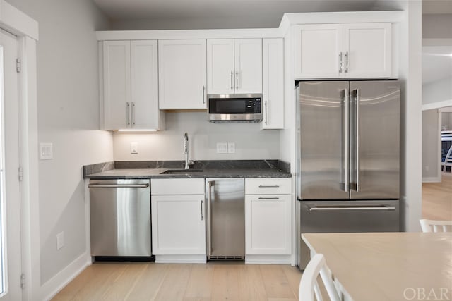 kitchen featuring light wood finished floors, appliances with stainless steel finishes, dark stone countertops, white cabinetry, and a sink