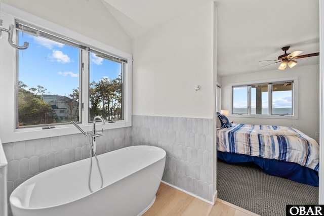 ensuite bathroom with a ceiling fan, wainscoting, lofted ceiling, a soaking tub, and wood finished floors