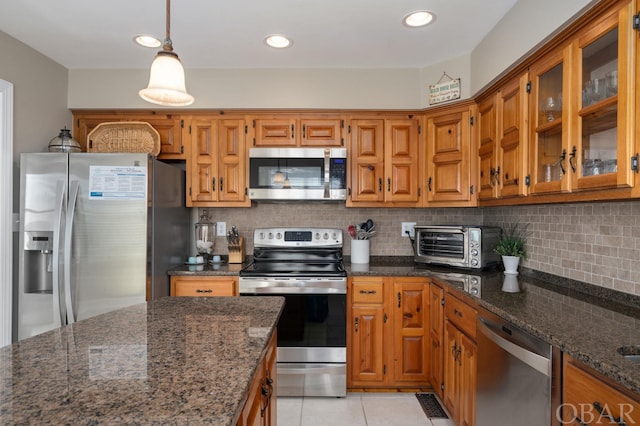 kitchen featuring pendant lighting, brown cabinets, stainless steel appliances, glass insert cabinets, and dark stone counters