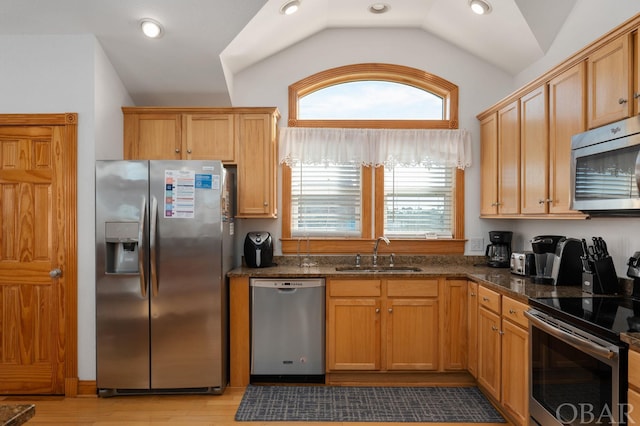 kitchen with appliances with stainless steel finishes, vaulted ceiling, a sink, dark stone countertops, and light wood-type flooring