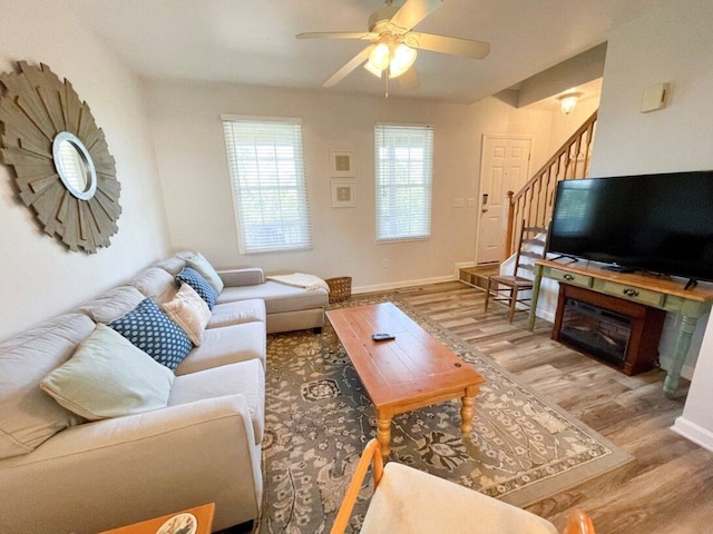 living area featuring baseboards, light wood-type flooring, ceiling fan, and stairway