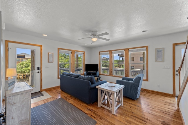 living area with a textured ceiling, ceiling fan, baseboards, stairway, and light wood-type flooring