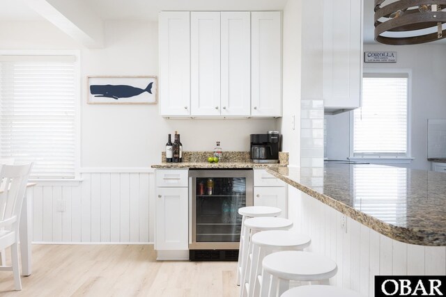 kitchen with beverage cooler, wainscoting, light wood-style flooring, light stone countertops, and white cabinetry