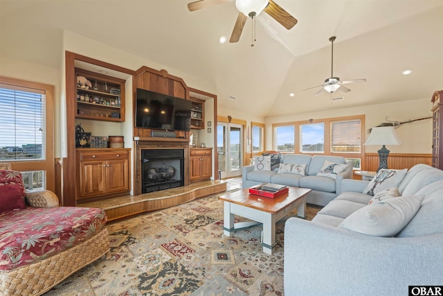 living area with lofted ceiling, a ceiling fan, a fireplace with raised hearth, and a wainscoted wall