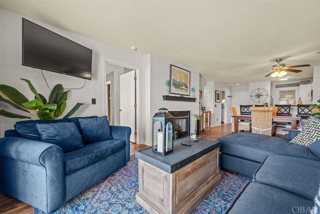 living area featuring dark wood-style floors, ceiling fan, a textured ceiling, and a glass covered fireplace