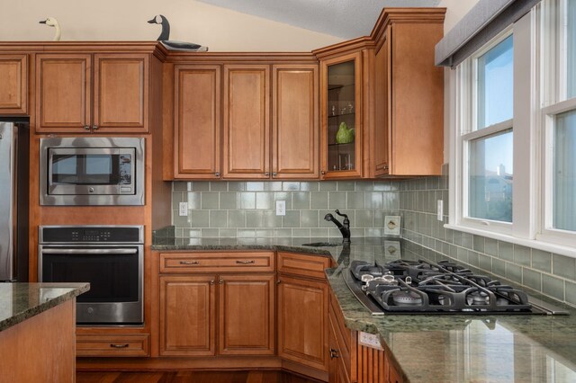 kitchen featuring a sink, appliances with stainless steel finishes, decorative backsplash, brown cabinetry, and dark stone countertops